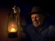 Old man holding lantern in dark room.